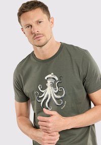 T-shirt vert olive en coton, featuring a graphique d'un poulpe blanc avec un chapeau de capitaine. Encolure ronde et manches courtes.