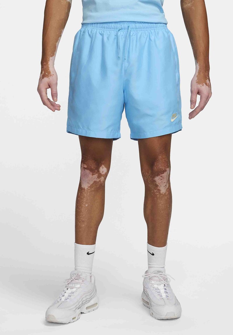 Person klädd i ljusblå Nike-shorts, vita Nike-strumpor och vita sneakers, stående mot en neutral bakgrund.