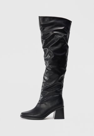 Botte en cuir noire montante jusqu'au genou avec tige détendue et talon bloc épais, vue de profil sur fond gris clair.