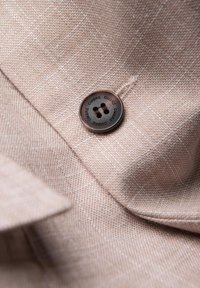 Gros plan sur un tissu beige texturé avec un bouton marron cousu, bouton gravé du mot « OMBRE » répété autour de son bord.