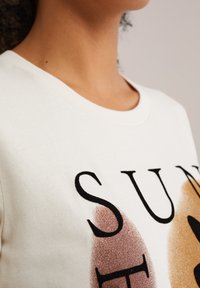 T-shirt en coton blanc avec un col rond, présentant du texte noir et deux taches de couleur abstraite en bronze et marron clair.