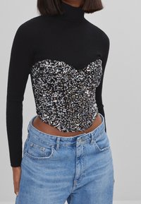 Svart, ribbad långärmad topp med paljettprydd byst och hjärtformad halsringning, kombinerad med ljusblå höga denimjeans.