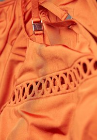 Tissu orange avec une texture lisse, surmonté d'un motif découpé décoratif le long de la couture et de quincaillerie pour sangle réglable.