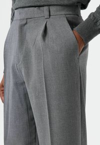 Pantalon gris sur mesure avec une texture subtile, doté de plis à l'avant et de poches latérales. La ceinture comporte des passants pour ceinture pour plus de fonctionnalité.