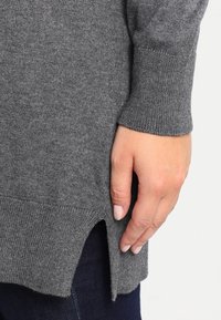 Szary dzianinowy sweter o prążkowanej fakturze, z bocznym rozcięciem i długimi rękawami, pokazany z ręką opartą na dolnym brzegu.