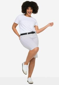 T-shirt blanc, shorts gris clair, ceinture tressée noire et baskets blanches avec des accents gris. Le modèle adopte une pose ludique, mettant en valeur sa tenue.