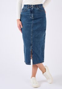 Denim-Midirock in Dunkelblau mit Frontschlitz, klassischem Fünf-Taschen-Design und sichtbaren Nahtdetails. Kombiniert mit weißen Sneakers.