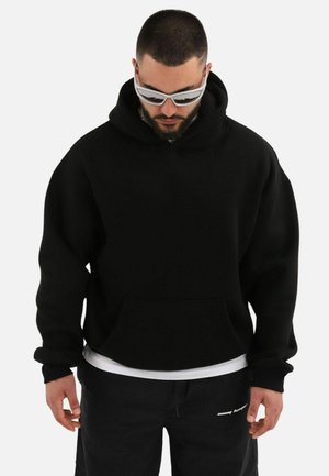 Black hoodie with a large front pocket, adjustable hood, and ribbed cuffs. Worn over a white shirt, paired with black pants and sunglasses.