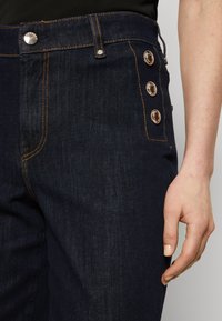 Jeans in denim blu scuro con cuciture in rame, vita alta e bottoni decorativi lungo il lato per un tocco di design aggiuntivo.