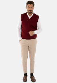 Gilet rosso in maglia sopra una camicia bianca con colletto, pantaloni beige e scarpe marroni. Il tessuto è liscio, con design a V e senza motivi visibili.