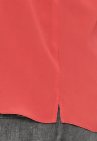 Coral blouse made of smooth fabric, featuring a curved hemline with side slits and stitching accents. The design is lightweight and breathable.