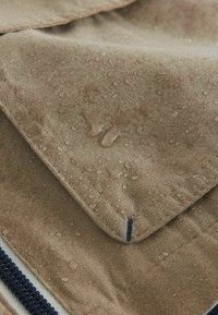 Gros plan sur un tissu kaki avec des gouttes d'eau perlées à la surface, montrant une texture résistante à l'eau et un détail de couture près d'une fermeture éclair.