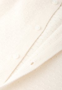Cárdigan de punto en color crema con un patrón texturizado. Presenta botones blancos redondos y suaves, y un diseño de cuello clásico. Fabricado con una tela suave y flexible.