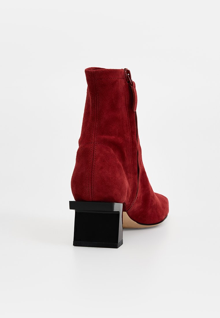 Czerwone zamszowe botki za kostkę na słupku, z bocznym zamkiem błyskawicznym i gładką teksturą. Obcas ma czarny akcent dla kontrastu.