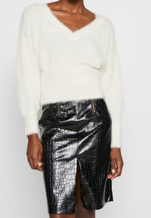 Femme portant un pull à col en V blanc duveteux et une jupe en cuir noir à texture crocodile avec une fente frontale et des détails zippés.