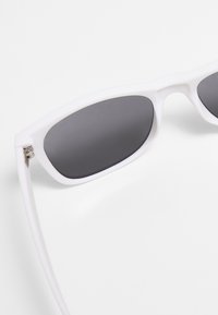 Ochelari de soare din plastic alb cu lentile gri, având un design geometric al ramelor și o finisare mată netedă. Bratele sunt subțiri și drepte.