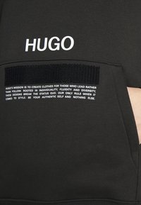 Zwarte sweatshirt met een wit "HUGO" logo op de borst en tekstdetails op de voorkantzak. Het materiaal lijkt zacht met een gladde textuur.