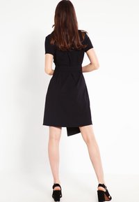 Mustapaitainen lyhythihainen mekko, jossa on istuva vyötärö, pleatattu selkäosa ja polven ylle ulottuva helma. Yhdistetty mustien korkokenkien kanssa.