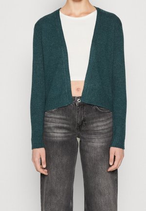 Personne portant un cardigan vert foncé ouvert devant, un top court blanc et un jean noir délavé taille haute.