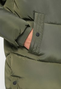 Pufferjakke i olivengrønn, med en glatt, blank tekstur, en hånd i en sidelomme og detaljer med ribbekanter.