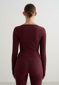 Burgunderrotes Langarmoberteil mit tailliertem Schnitt und gerippter Textur, kombiniert mit passenden gerippten Leggings, von hinten betrachtet.
