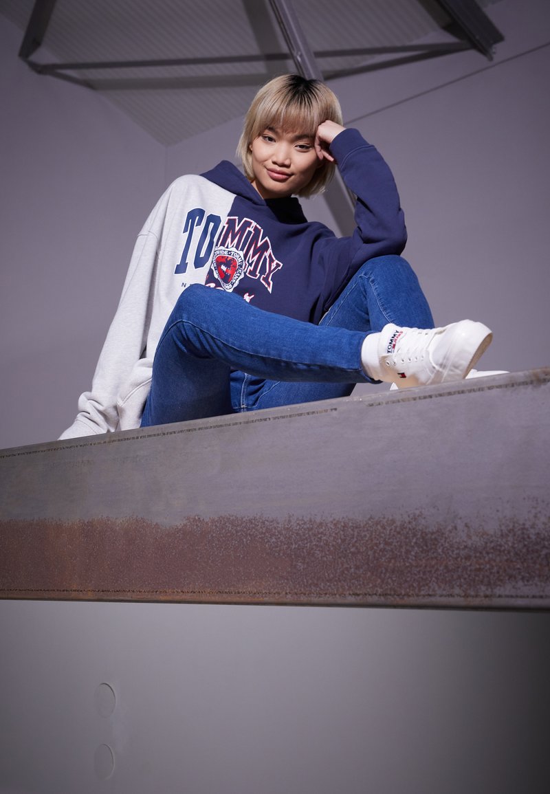 Felpa con cappuccio grigia e blu navy, con il logo "TOMMY", abbinata a jeans blu attillati e sneakers bianche, su uno sfondo neutro.