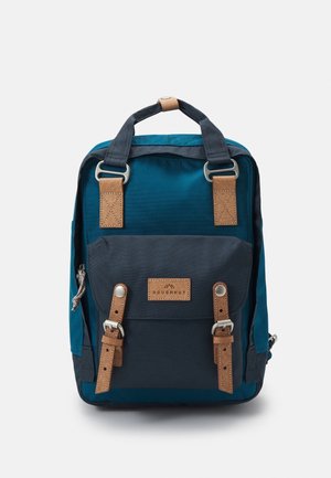 Zaino blu e navy con cinghie in pelle marrone e fibbie in metallo, tasca frontale e maniglie superiori per il trasporto su sfondo bianco.