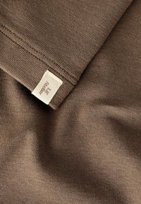 Brązowa dzianina o gładkiej fakturze, z małą metką w rogu. Tkanina miękko się układa, ukazując drobne prążkowanie.