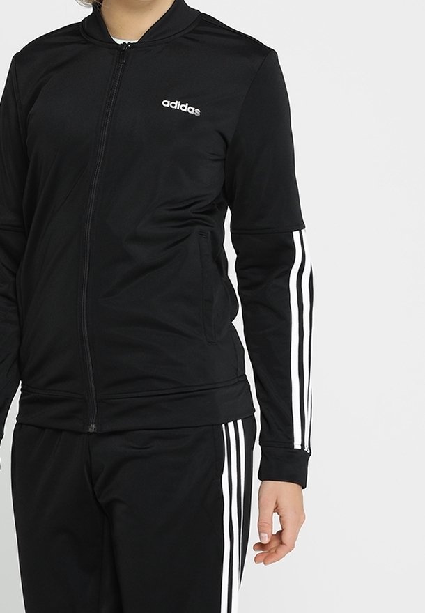 Personne portant un survêtement Adidas noir avec des bandes blanches sur les manches et le pantalon, veste zippée et main visible sur le côté.