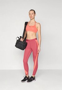 Ryškiai koraliniam atletiškam liemenėlė PUMA, tamsiai raudonos spalvos aukšto juosmens tamprės su rožiniais akcentais ir juoda sporto krepšys, derinami su juodais sportbačiais.