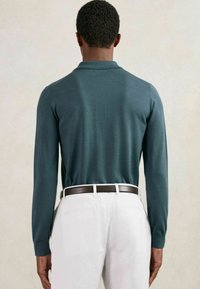 Cămașă cu mâneci lungi și guler din material tricotat de culoare verde închis; textură fină, asortată cu pantaloni albi și o curea maro, văzută din spate.