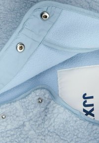 Tissu polaire bleu clair avec une surface douce et texturée ; comporte des boutons-pressions argentés et une étiquette blanche avec texte bleu.