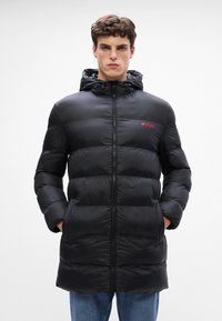 Chaqueta acolchada negra con relleno horizontal, cierre de cremallera y capucha ajustable. Presenta un pequeño logo rojo de "HUGO" en el pecho.