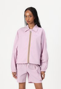 Lichtroze windjack met een kraag en elastische manchetten, voorzien van een ritssluiting aan de voorkant met gele accenten, gecombineerd met bijpassende shorts.