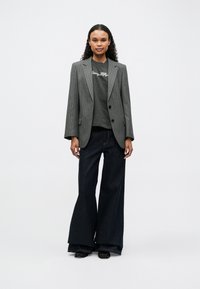 Blazer gris a rayas finas sobre una camisa gris oscura, combinado con unos vaqueros oscuros de pernera ancha. Calzado negro con un acabado elegante.