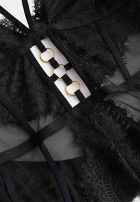 Průhledné černé spodní prádlo s krajkovými lemy, saténovými ramínky, trojúhelníkovými výřezy a zlatými kroužky ve středu přední části pro důraz.