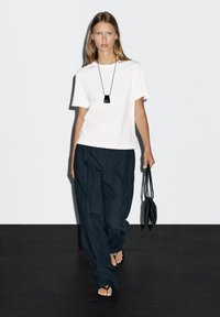 Maglietta bianca in cotone, pantaloni larghi a righe blu navy, sandali neri con cinturini incrociati e una collana nera con ciondolo, con una piccola borsa nera in mano.