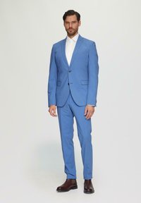 Lichtblauw op maat gemaakt pak met een enkele knoopjasje, slim-fit broek en een witte blouse. Bruine leren laarzen maken de outfit compleet.