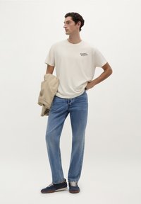 Beige bomulds t-shirt med logo, lyseblå jeans, tan jakke hængende over armen, og marineblå sneakers med grå snørebånd og brune såler.