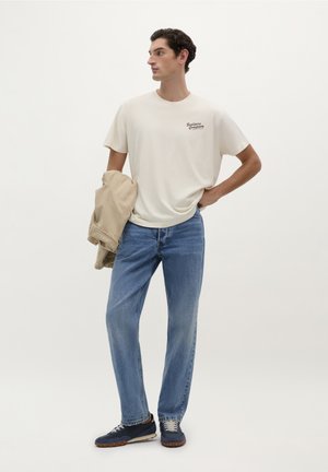 T-shirt in cotone beige con logo, jeans azzurri chiari, giacca color cammello appoggiata sul braccio e sneakers navy con lacci grigi e suola marrone.