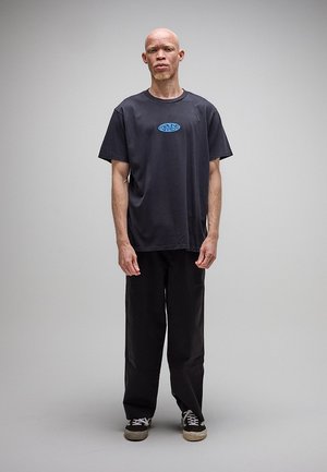 Man met albinisme staat neutraal, draagt een zwart T-shirt met een blauw logo, zwarte broek en zwarte sneakers met witte veters tegen een effen grijze achtergrond.