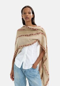 Beżowy, spleciony szalik z czerwonymi wzorami geometrycznymi i paskami. Miękka tekstura, zarzucony na ramiona, uzupełniający białą koszulę i niebieskie dżinsy.