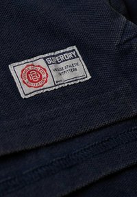 Marineblaue, strukturierte Stoff mit einem rechteckigen weißen Etikett, das roten und blauen Text trägt und "Superdry Vintage Athletic Outfitters" liest.