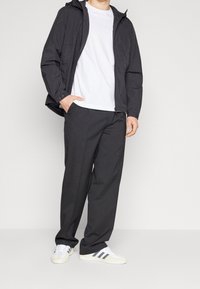 Person iført sort hættejakke, hvid t-shirt, løse sorte bukser og hvide sneakers med sorte striber, står med den ene hånd i lommen.