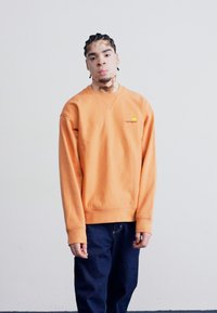 Orange bomulds sweatshirt med rund halsudskæring og ribbede manchetter, med et lille broderet logo. Båret med mørkeblå jeans.