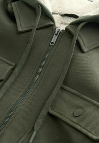 Artimiausio plano alyvuogių žalios striukės su užtrauktuku, raišteliais, snapiniais užsegimo kišenės antklodė ir matoma pūkuota balta pamušalu.