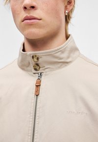 Μπεζ ελαφρύ μπουφάν με ψηλό γιακά, δύο κουμπιά και κλείσιμο με φερμουάρ. Διαθέτει ανάγλυφο λογότυπο και δερμάτινο λουράκι ως διακοσμητικό. Ομαλή υφή.