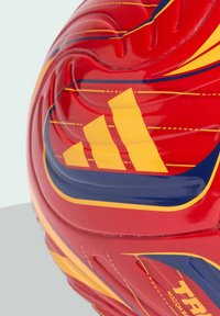 Zatvoren kadar crvene Adidas lopte za nogomet s žutim i tamnoplavim prugama i reljefnim detaljima na svijetloj pozadini.