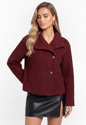 Frau mit langen, welligen blonden Haaren, die eine burgunderrote strukturierte Jacke mit Knöpfen und einen schwarzen Leder-Minirock mit seitlichem Schlitz trägt.