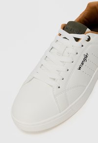 Baskets blanches avec une tige synthétique lisse, bout rond et lacets plats. Comprend une languette grise avec un logo et des perforations sur les côtés pour la ventilation.